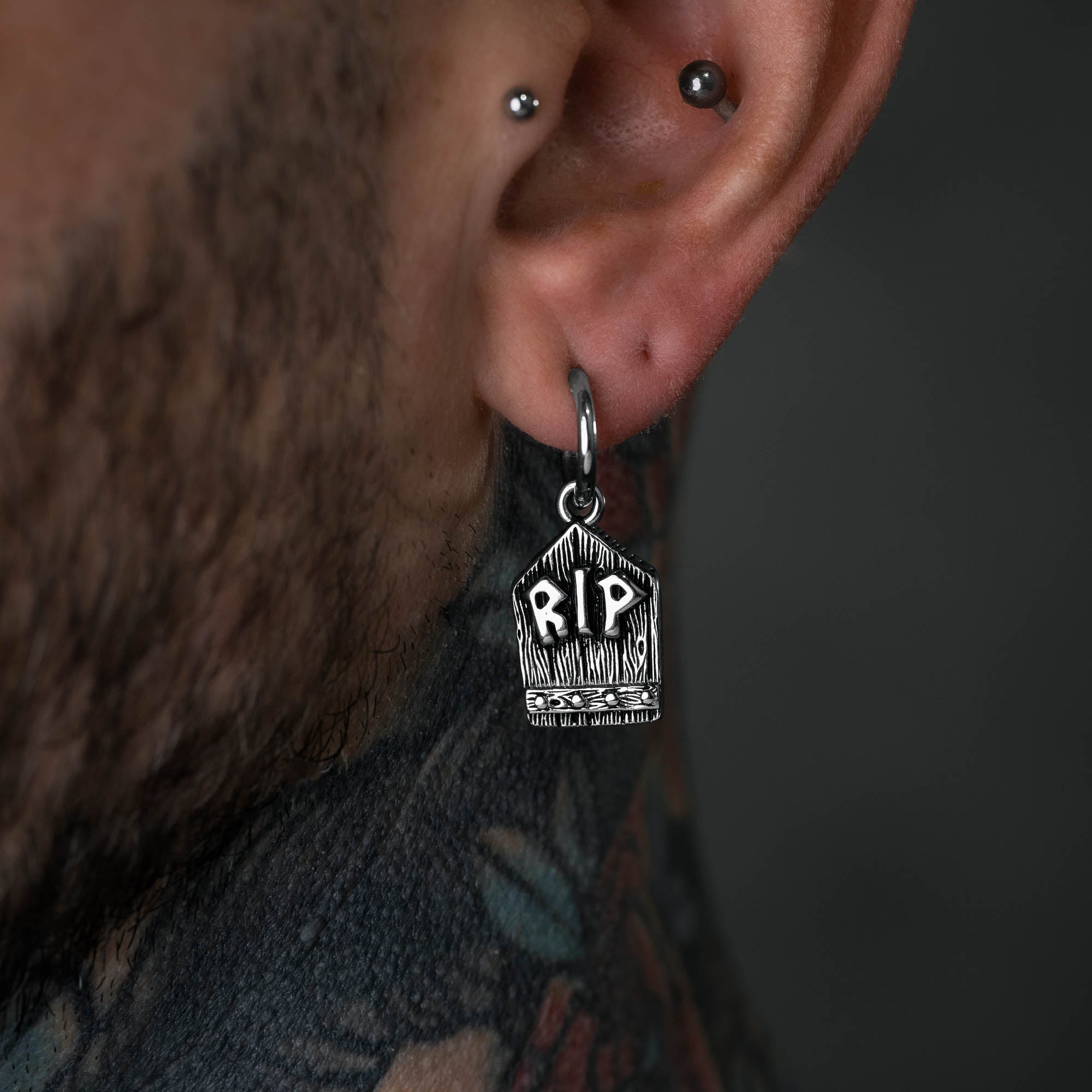 Silver grave stone earring with "RIP" embedded on male model with neck tattoos