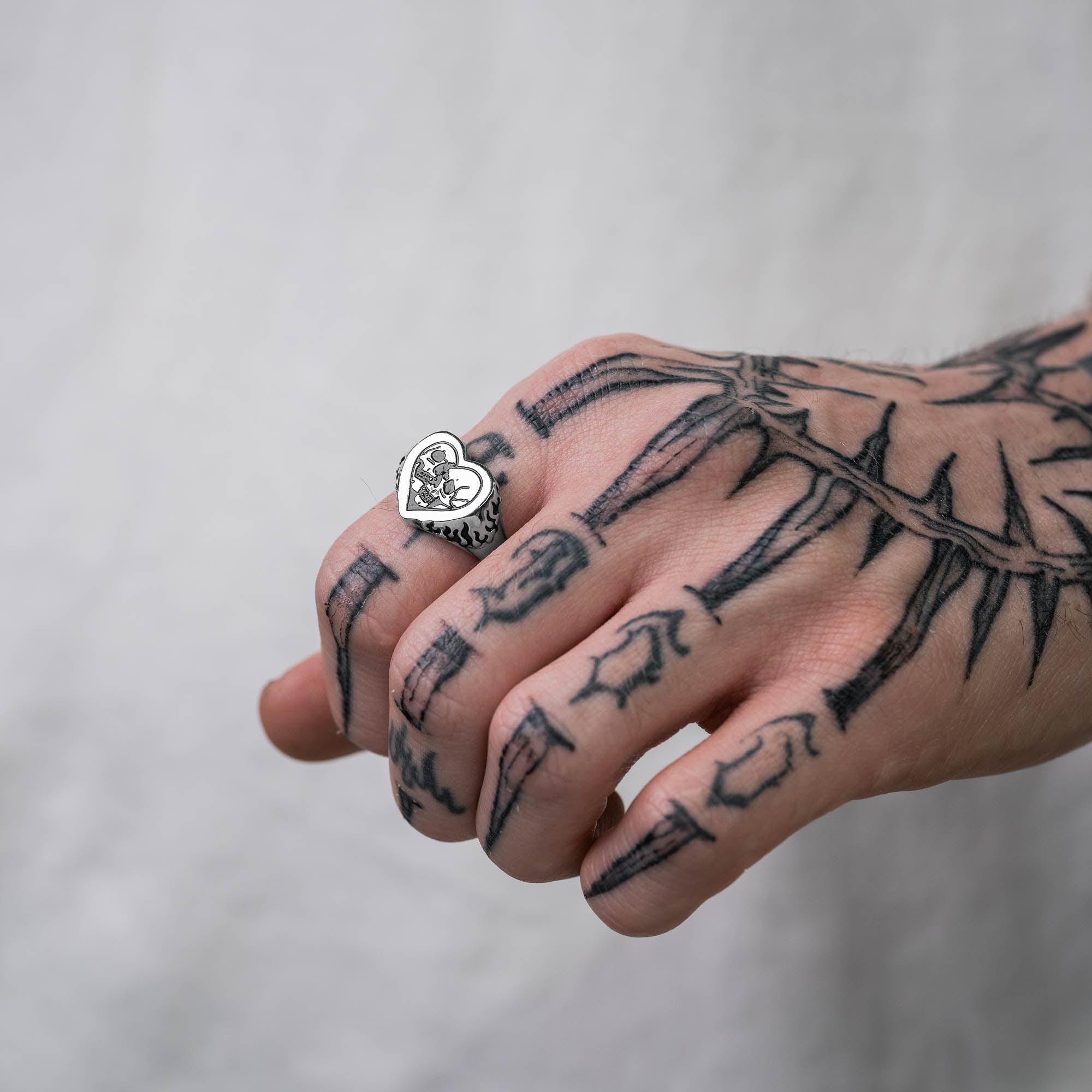 Silver heart shaped ring with skull details on ring face and flame details on sides, on male hand with tattoos