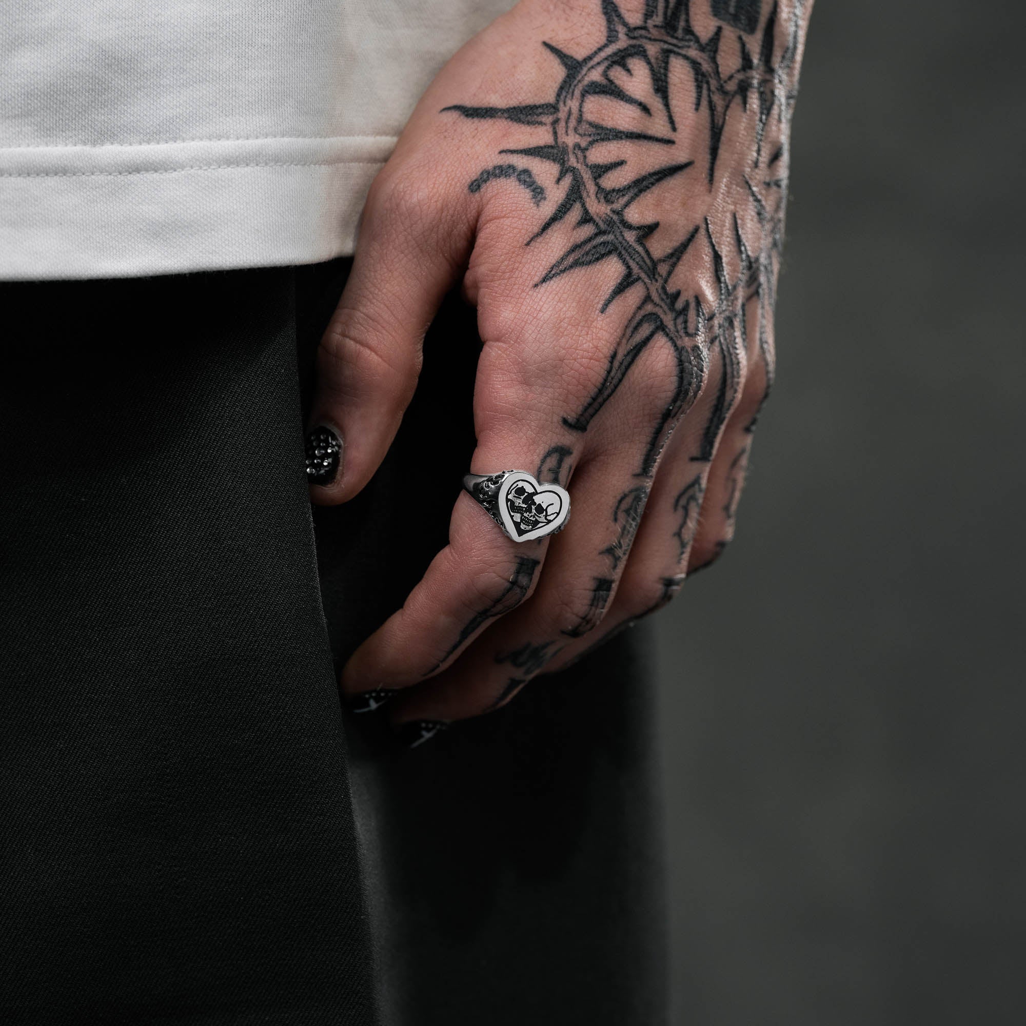 Silver heart shaped ring with skull details on ring face and flame details on sides, on male hand with tattoos