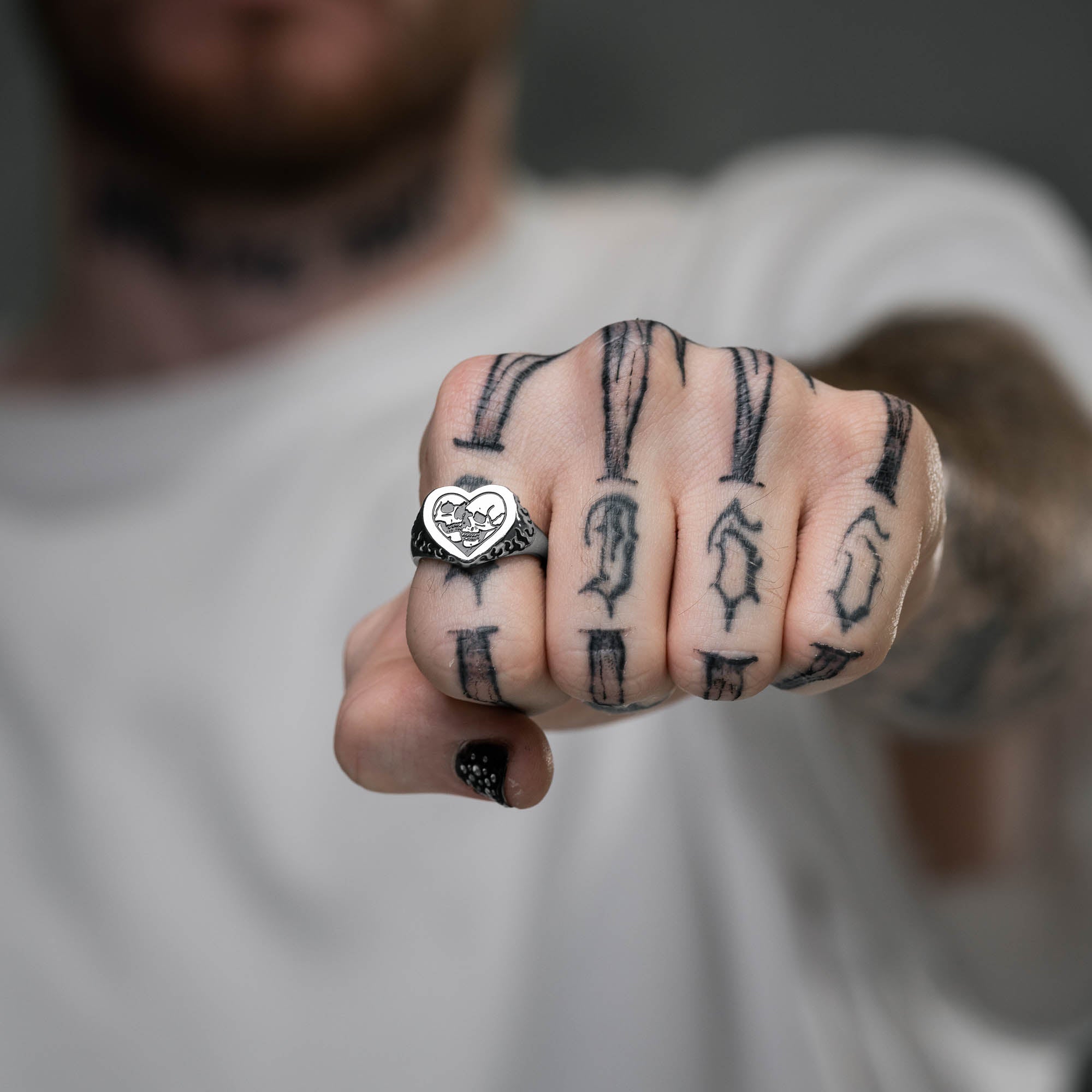 Silver heart shaped ring with skull details on ring face and flame details on sides, on male hand with tattoos