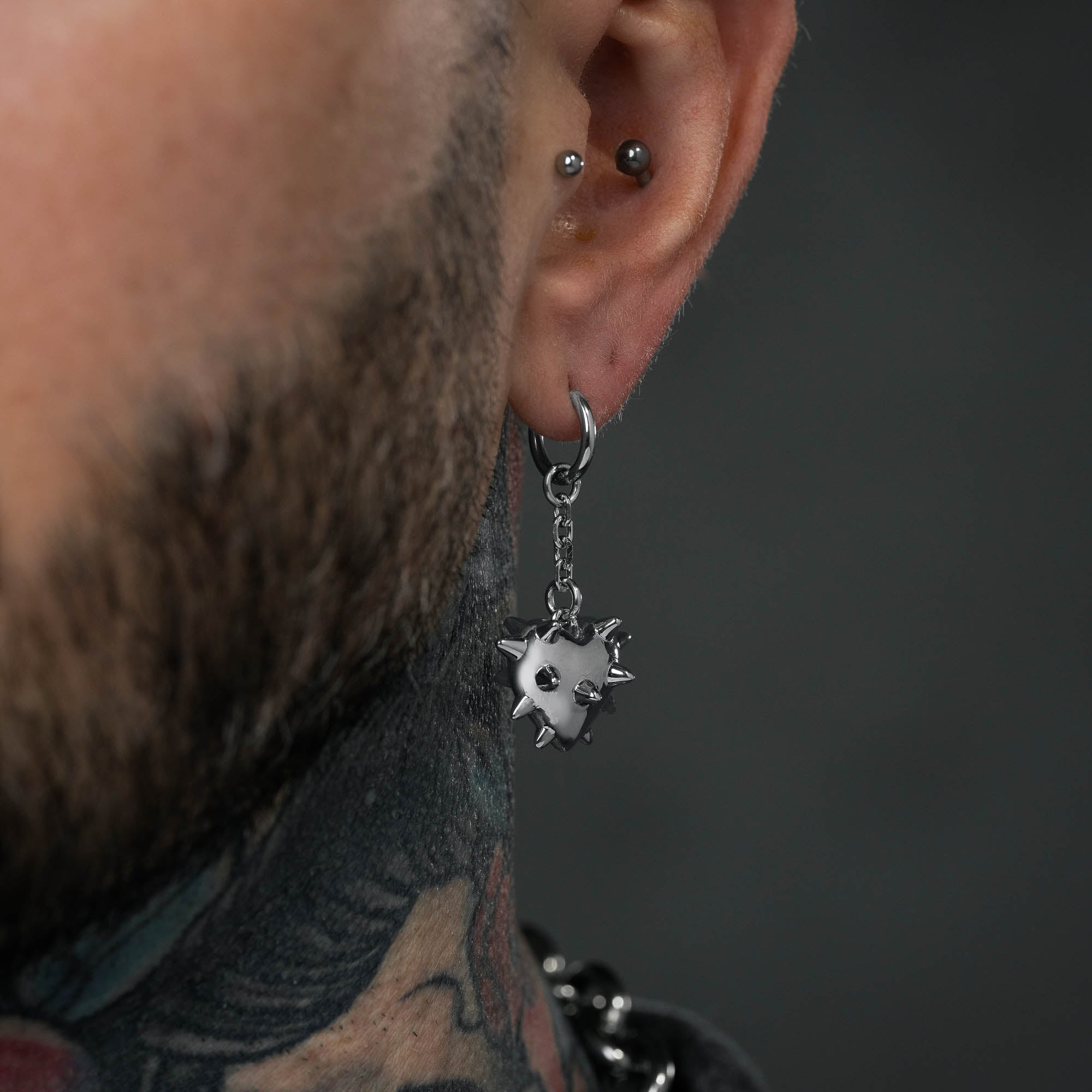 Silver dangling earrings with spiked love heart hanging from chain, on male model with neck tattoos.