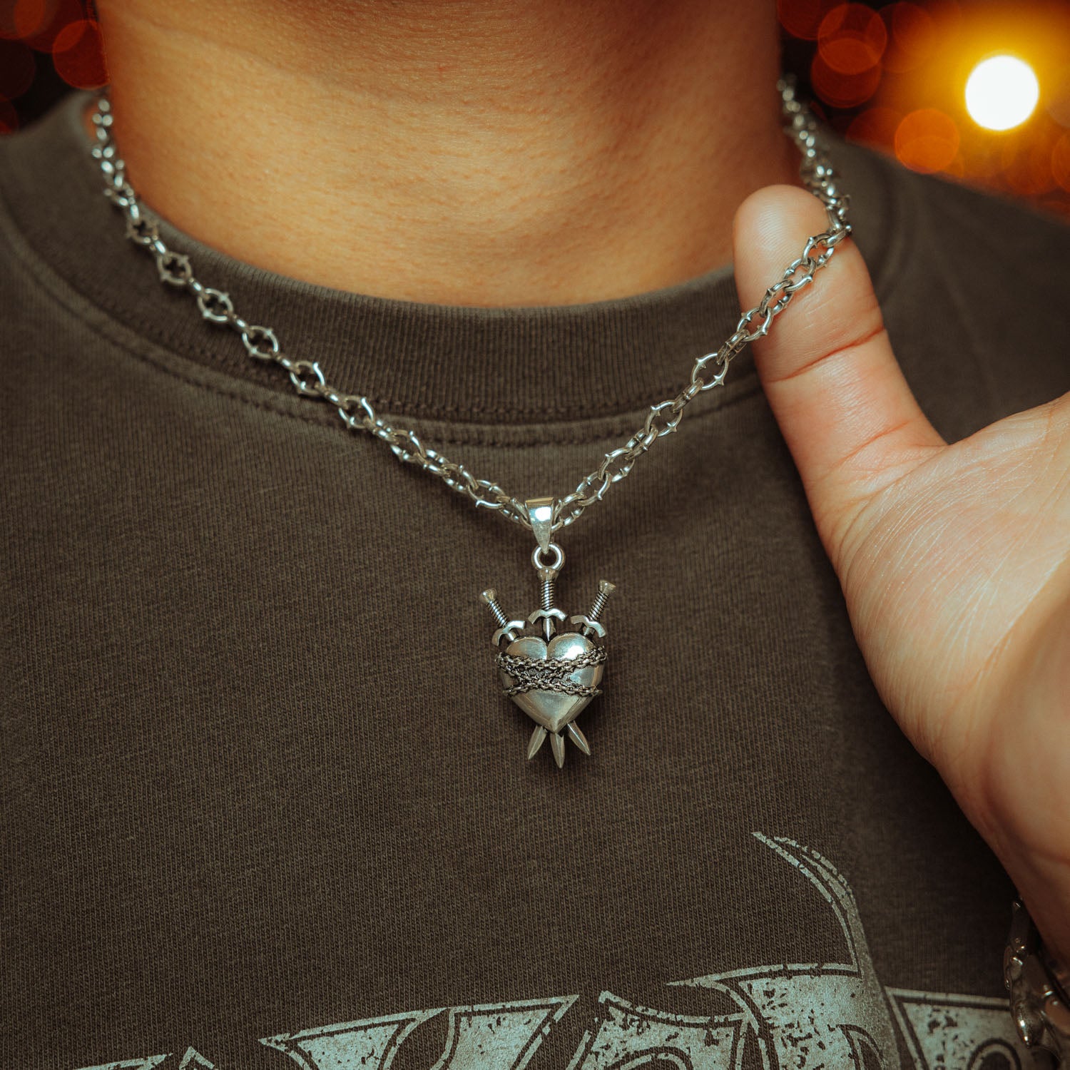 Silver heart pendant with three swords going through it and chain wrapped around it on a spiked chain on a male model with grey shirt